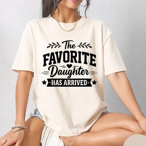 May include: Ivory t-shirt featuring the text "The Favorite Daughter Has Arrived" in black. The design includes leaves, hearts, and stars. The shirt is a casual style. The colour is described as ivory.