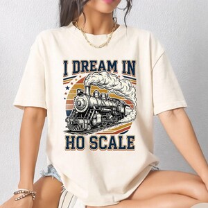 Può includere: T-shirt color crema con grafica di un treno vintage e la scritta "I Dream in HO Scale". Il design include una locomotiva a vapore, un tramonto e stelle. La maglietta è a maniche corte e girocollo.