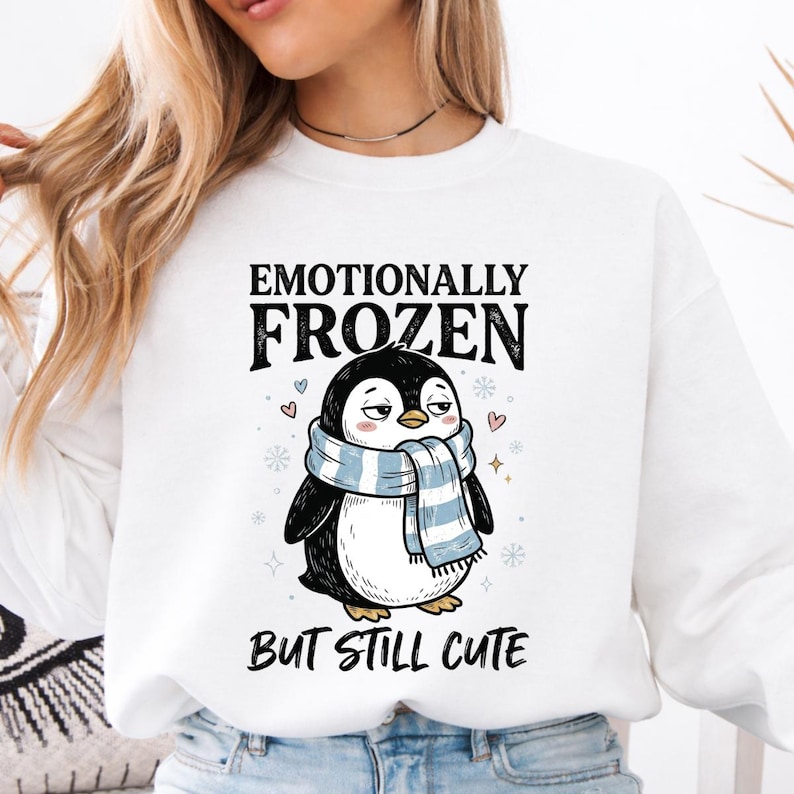 May include: White sweatshirt featuring a cartoon penguin wearing a blue and white striped scarf. The text above the penguin reads "Emotionally Frozen", and below it, "But Still Cute". The design includes hearts and snowflakes.