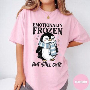 May include: A light pink t-shirt featuring a cartoon penguin wearing a blue and white scarf. The text above the penguin reads "Emotionally Frozen" and below it, "But Still Cute."