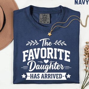 May include: Navy blue t-shirt with the text "The Favorite Daughter Has Arrived" in white. The shirt has a crew neck and short sleeves. A gold necklace with a star pendant is draped across the shirt.