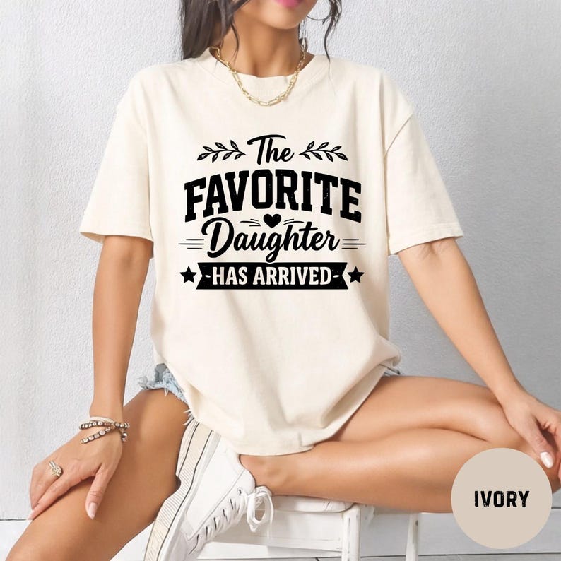 May include: Ivory t-shirt featuring the text "The Favorite Daughter Has Arrived" in black. The design includes leaves, hearts, and stars. The shirt is a casual style. The colour is described as ivory.