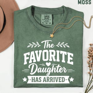 May include: A moss green t-shirt with the text "The Favorite Daughter Has Arrived" in white. A gold star pendant necklace and a brown hat are also visible. The shirt is a Comfort Colors XL.