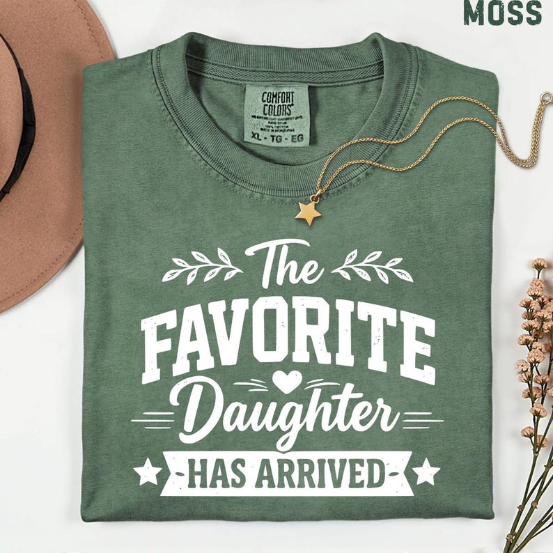 May include: A moss green t-shirt with the text "The Favorite Daughter Has Arrived" in white. A gold star pendant necklace and a brown hat are also visible. The shirt is a Comfort Colors XL.