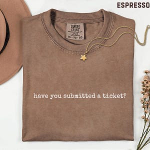May include: Espresso-colored t-shirt with the text "have you submitted a ticket?" in white. A gold star pendant necklace rests on the shirt. The shirt is folded and has a Comfort Colors tag.