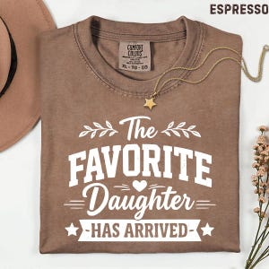 May include: Espresso-coloured t-shirt with the text "The Favorite Daughter Has Arrived" in white. A gold star pendant necklace and a brown hat are also visible. The shirt is a Comfort Colors XL-TG-EG.