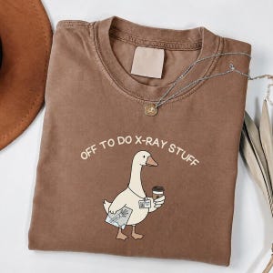 May include: A brown crewneck t-shirt featuring a graphic of a white goose wearing an ID badge, holding a coffee cup and an X-ray film. The text above the goose reads "OFF TO DO X-RAY STUFF" in a simple, light-colored, sans-serif font.