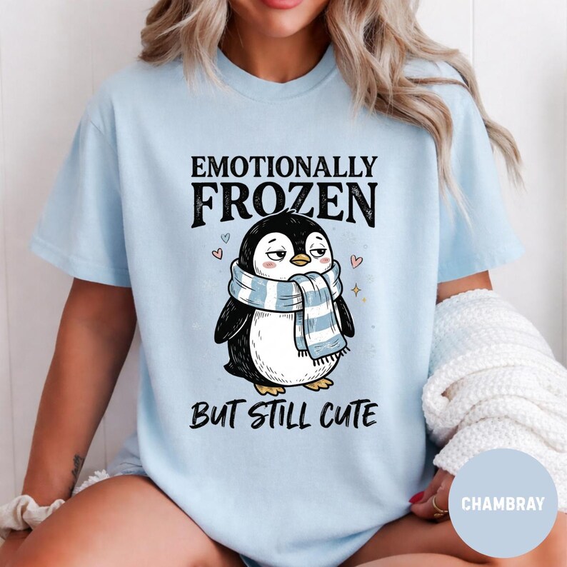 May include: Light blue t-shirt featuring a cartoon penguin wearing a blue and white scarf. The text on the shirt reads "Emotionally Frozen But Still Cute". The penguin has a slightly grumpy expression.