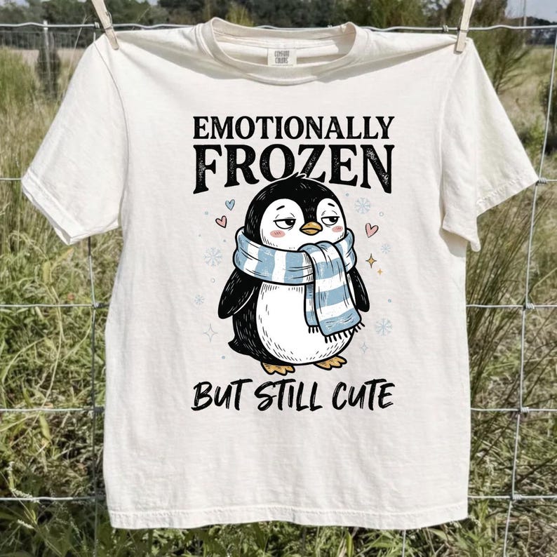 May include: Cream-colored t-shirt featuring a cartoon penguin wearing a blue and white scarf. The text above the penguin reads "Emotionally Frozen," and below it, "But Still Cute." The shirt is hanging on a clothesline.