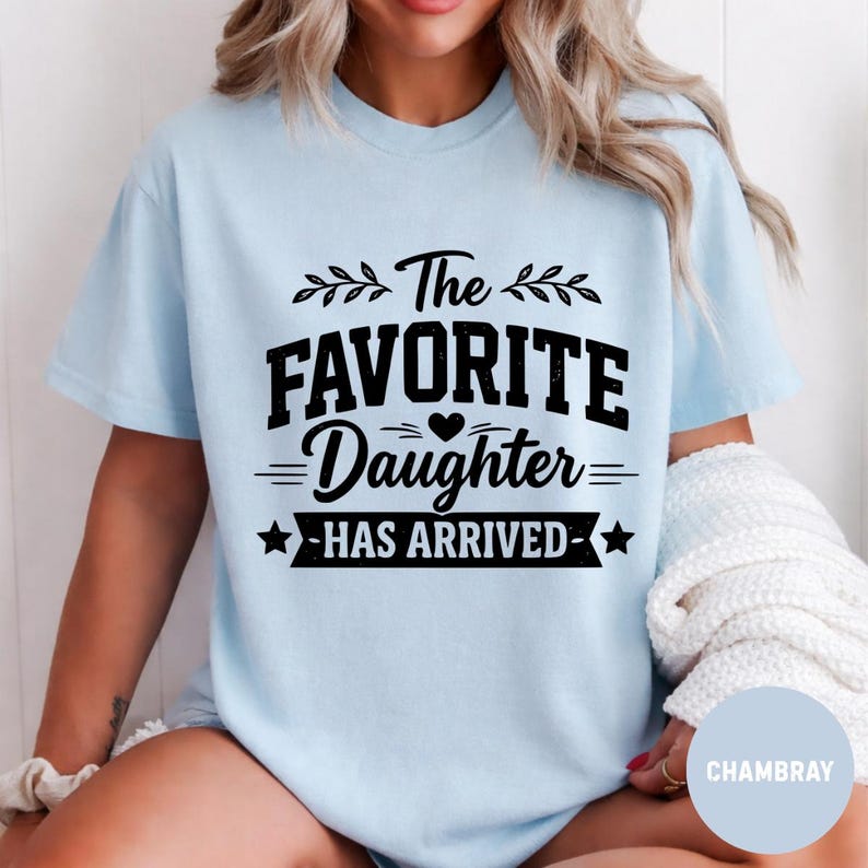 May include: Pale blue t-shirt featuring the black text "The Favorite Daughter Has Arrived". The design includes decorative leaves, a heart, and stars. The shirt is made of a soft material.