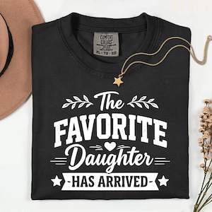 May include: Black crew neck t-shirt with the text "The Favorite Daughter Has Arrived" in white. A gold star pendant necklace and a brown hat are also visible. The shirt has a Comfort Colors tag.
