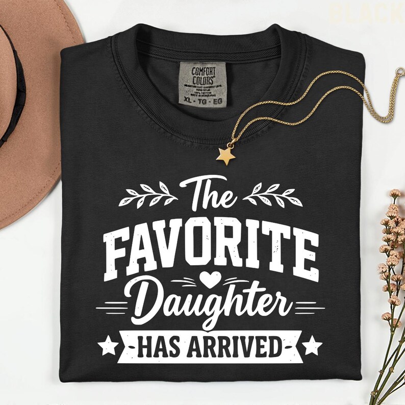 May include: Black crew neck t-shirt with the text "The Favorite Daughter Has Arrived" in white. A gold star pendant necklace and a brown hat are also visible. The shirt has a Comfort Colors tag.