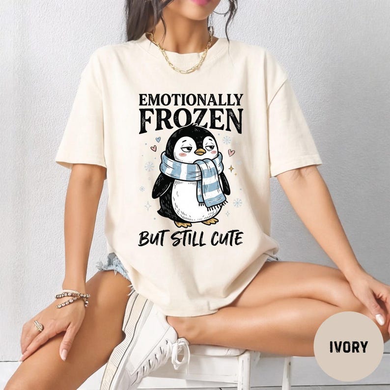 May include: Ivory-colored t-shirt featuring a cartoon penguin wearing a blue and white striped scarf. The text above the penguin reads "Emotionally Frozen" and below it, "But Still Cute". The shirt is a relaxed fit.