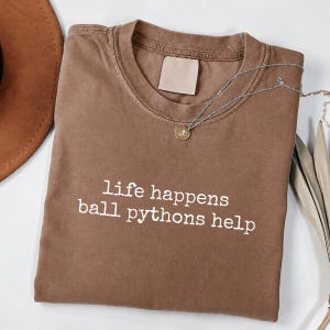 Puede incluir: Una camiseta marrón de manga corta con el texto blanco "life happens ball pythons help" impreso en la parte delantera. La prenda tiene un cuello redondo acanalado, una etiqueta rectangular en blanco en el interior y un collar plateado fino.