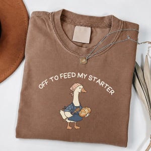 May include: A brown short-sleeved t-shirt featuring a graphic of a goose wearing a blue apron and a checkered headscarf, holding a wooden spoon and a loaf of bread. The text "OFF TO FEED MY STARTER" is printed in a white arc above the goose illustration.
