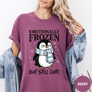 May include: Berry-colored t-shirt featuring a cartoon penguin wearing a blue and white scarf. The text on the shirt reads "Emotionally Frozen But Still Cute". The design includes snowflakes and stars.