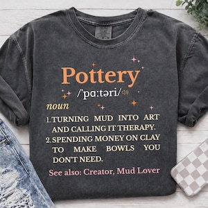 May include: Dark gray t-shirt with the word "Pottery" in orange, along with its pronunciation and definition. The definition includes phrases like "turning mud into art" and "spending money on clay." The shirt also lists "Creator, Mud Lover."