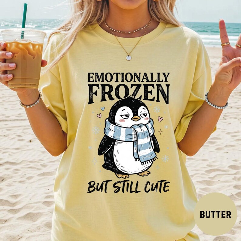 May include: Butter-colored t-shirt with the text "Emotionally Frozen But Still Cute" and a cartoon penguin wearing a blue and white scarf. The penguin has a slightly grumpy expression. The shirt is a casual style.