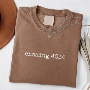 May include: A brown crew neck t-shirt featuring the white text "chasing 4014" printed across the chest. The garment has a ribbed collar and a blank rectangular label inside the neckline, accompanied by a delicate gold-toned chain necklace.