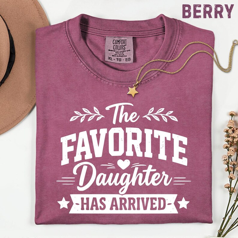 May include: A berry-coloured t-shirt with the text "The Favorite Daughter Has Arrived" in white. A gold-coloured star pendant necklace is draped across the shirt. The shirt is a Comfort Colors brand, size XL-TG-EG.
