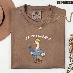 以下が含まれることがあります： 「OFF TO ENGINEER」の文字と、ヘルメットをかぶり設計図を持つ漫画のガチョウが描かれた、エスプレッソ色のTシャツ。金の星のペンダントネックレスがシャツにかけられています。シャツにはXL-TG-EGと記載されています。