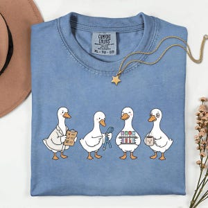 Op de afbeelding: Een blauw t-shirt met een afbeelding van vier witte eenden verkleed als medische professionals. Eén eend houdt een klembord vast, één een blauw lint, één een reageerbuisrekje en één een koffiemok. Het shirt heeft een Comfort Colors label.