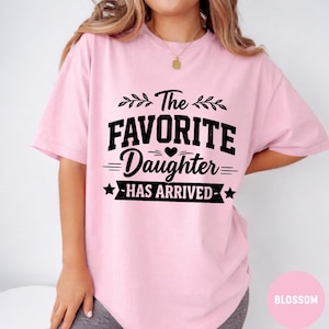May include: A pale pink t-shirt featuring the black text "The Favorite Daughter Has Arrived". The text is in a bold, stylized font with decorative elements. The shirt is made of a soft, comfortable material.