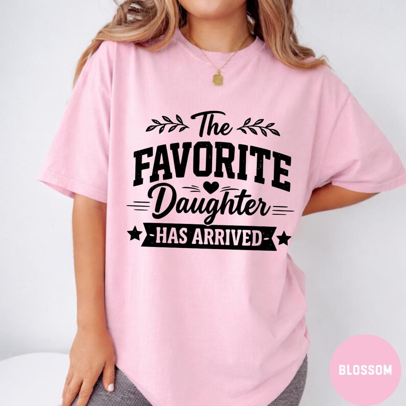 May include: A pale pink t-shirt featuring the black text "The Favorite Daughter Has Arrived". The text is in a bold, stylized font with decorative elements. The shirt is made of a soft, comfortable material.