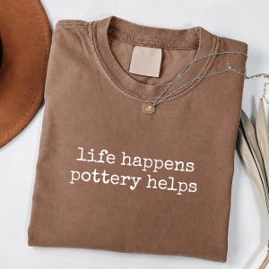 Può includere: Una t-shirt marrone a maniche corte con scollo rotondo e un'etichetta rettangolare vuota all'interno del colletto. La parte anteriore presenta il testo bianco "life happens pottery helps" stampato con un carattere in stile macchina da scrivere.