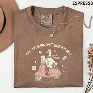 May include: Espresso-colored t-shirt with the phrase "OFF TO IMPROVE BREATHING" and a cartoon duck riding a pink scooter. The shirt has a crew neck and a Comfort Colors tag. A gold necklace with a star pendant is draped across the shirt.