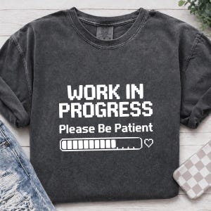 May include: Dark gray long-sleeve t-shirt with the text "WORK IN PROGRESS Please Be Patient" in white block letters. A loading bar graphic and a heart symbol are below the text. The shirt is made of a soft, comfortable material.