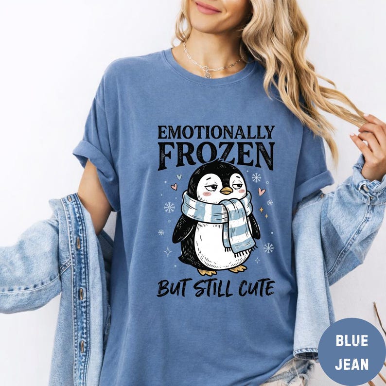 May include: A blue t-shirt with the text "Emotionally Frozen But Still Cute" above a cartoon penguin wearing a blue and white scarf. The penguin is black and white with a slightly grumpy expression. The shirt has rolled-up sleeves.