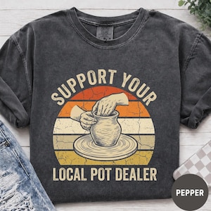 May include: A charcoal grey t-shirt with a graphic print featuring a circular sunset background in orange and yellow. The design shows hands shaping a clay pot on a wheel with the text SUPPORT YOUR LOCAL POT DEALER in a bold, arched font.