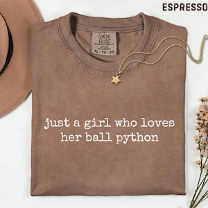 May include: An espresso-colored t-shirt featuring the white text "just a girl who loves her ball python" printed on the front. The shirt has a Comfort Colors brand label at the inner neckline and is paired with a gold star pendant necklace.