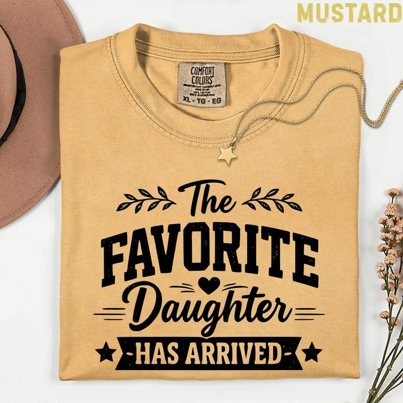 May include: Mustard-coloured t-shirt with the text "The Favorite Daughter Has Arrived" in black. The shirt has a crew neck and short sleeves. A gold necklace with a star pendant is on the shirt. A brown hat and flowers are in the background.