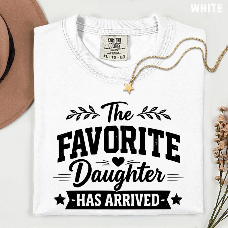 May include: A white t-shirt with the text "The Favorite Daughter Has Arrived" in black. The shirt has a crew neck and a tag that says "Comfort Colors". A gold necklace with a star pendant is draped on the shirt.