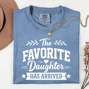 May include: A light blue Comfort Colors t-shirt with the text "The Favorite Daughter Has Arrived" in white. A gold star pendant necklace and a brown hat are also visible. The shirt is XL-TG-EG.