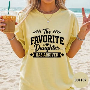 May include: Pale yellow t-shirt featuring the black text "The FAVORITE Daughter HAS ARRIVED". The design includes a heart and stars. The shirt is a casual style.