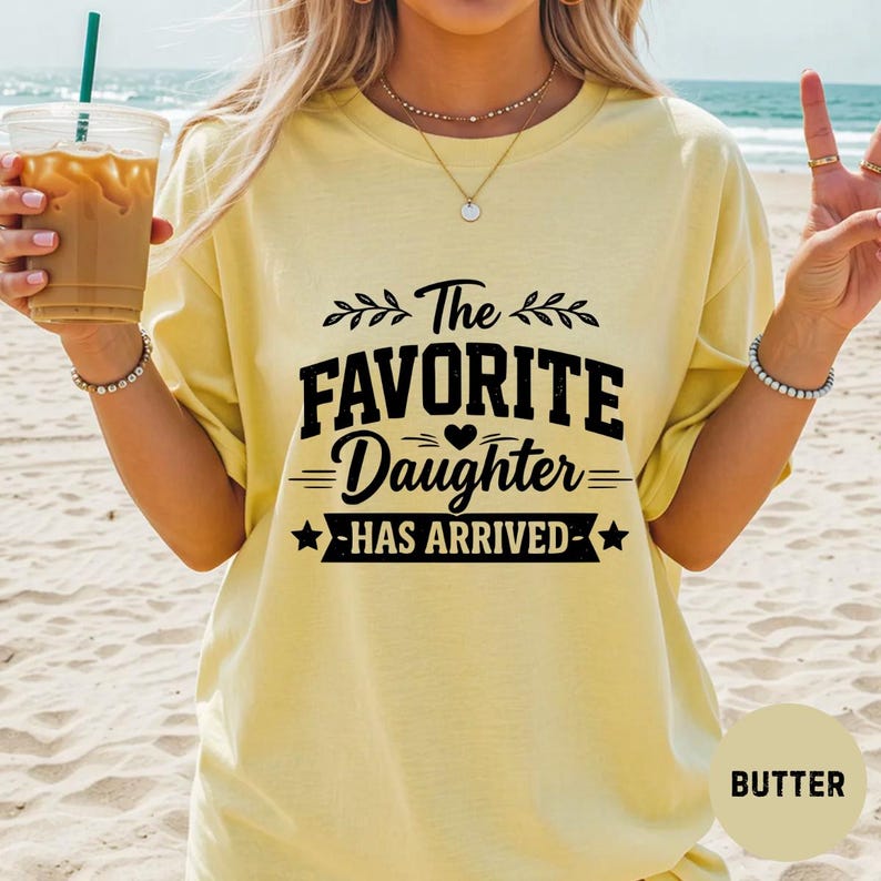 May include: Pale yellow t-shirt featuring the black text "The FAVORITE Daughter HAS ARRIVED". The design includes a heart and stars. The shirt is a casual style.