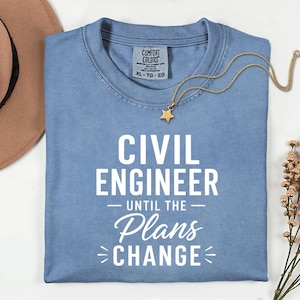 May include: A light blue t-shirt with the text "CIVIL ENGINEER - UNTIL THE Plans CHANGE" in white. A gold necklace with a star pendant rests on the shirt. A brown hat and sprig of flowers are in the background.