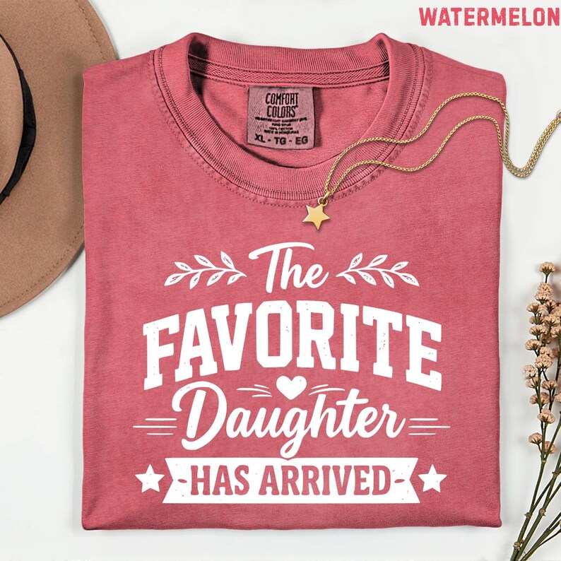 May include: A crimson t-shirt with the white text "The Favorite Daughter Has Arrived". A gold necklace with a star pendant lies on the folded shirt. The shirt has a Comfort Colors label. The word "WATERMELON" is at the top.