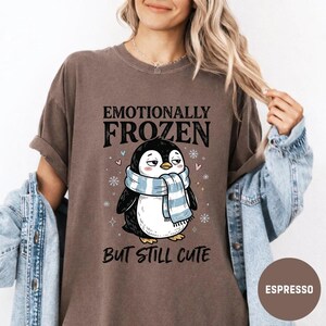 May include: Espresso-colored t-shirt featuring a cartoon penguin wearing a blue and white scarf. The text "Emotionally Frozen But Still Cute" is printed above and below the penguin illustration. The shirt has a relaxed fit.
