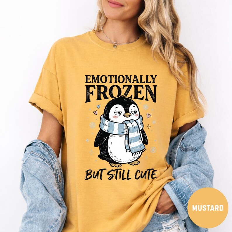 May include: Mustard-colored t-shirt with the text "Emotionally Frozen But Still Cute" and a cartoon penguin wearing a blue and white scarf. The penguin has a slightly grumpy expression. The shirt is a relaxed fit.
