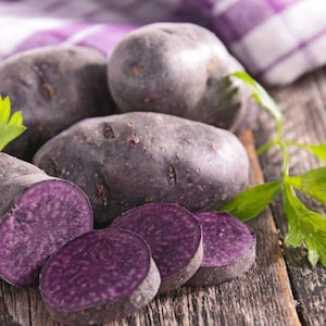 May include: Several whole purple potatoes and three sliced purple potato rounds arranged together. The potatoes have dark purple skin and deep purple flesh. Fresh green parsley leaves are placed near the sliced potato pieces.