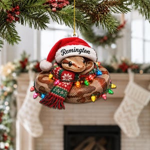 Op de afbeelding: Een ronde kerstversiering met een opgerolde bruine slang die een rood-witte kerstmuts draagt met de naam Remington op de rand. De slang is gewikkeld in een rood, groen en wit sjaaltje met patronen en kleurrijke kerstverlichting.