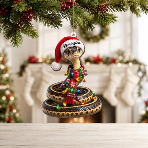 Könnte beinhalten: Ein flacher Hängeschmuck in Form einer zusammengerollten Schlange mit schwarzen und gelben Schuppen. Die Schlange trägt eine rot-weiße Weihnachtsmütze mit dem Namen Remington auf dem Rand und einen rot-grün-weiß gemusterten Schal mit Lichterkette.