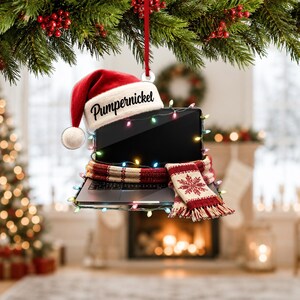 Peut inclure: Une décoration de Noël plate en forme d'ordinateur portable portant un bonnet de Père Noël rouge et blanc avec le nom Pumpernickel imprimé sur le bord. L'ordinateur est enveloppé dans une écharpe en tricot rouge et blanc avec des guirlandes.