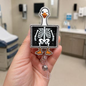 May include: A badge reel featuring a white goose with an orange beak and feet. The goose is positioned in front of a square black frame displaying a white skeletal X-ray of a ribcage and pelvis. The entire design has a silver glitter border.