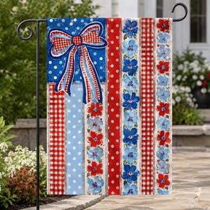 May include: A patriotic garden flag with a bow and stripes in red, white, and blue. The flag features a large bow on a blue polka-dot background, with alternating stripes of red polka dots, gingham, and floral patterns. The flag is displayed on a black metal stand.