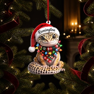 May include: A leopard gecko Christmas ornament featuring the name Remington in black script on a red and white Santa hat. The gecko wears a green, red, and white patterned scarf wrapped in colourful, glowing string lights, hanging by a red ribbon.